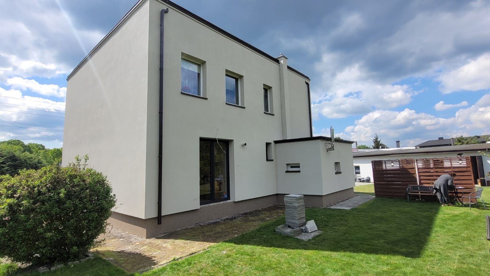 Elewacja domu po tynkowaniu i malowaniu na jasny kolor. Widoczny fragment ogrodu, okna z czarnymi ramami i rury spustowe. Słoneczny dzień.