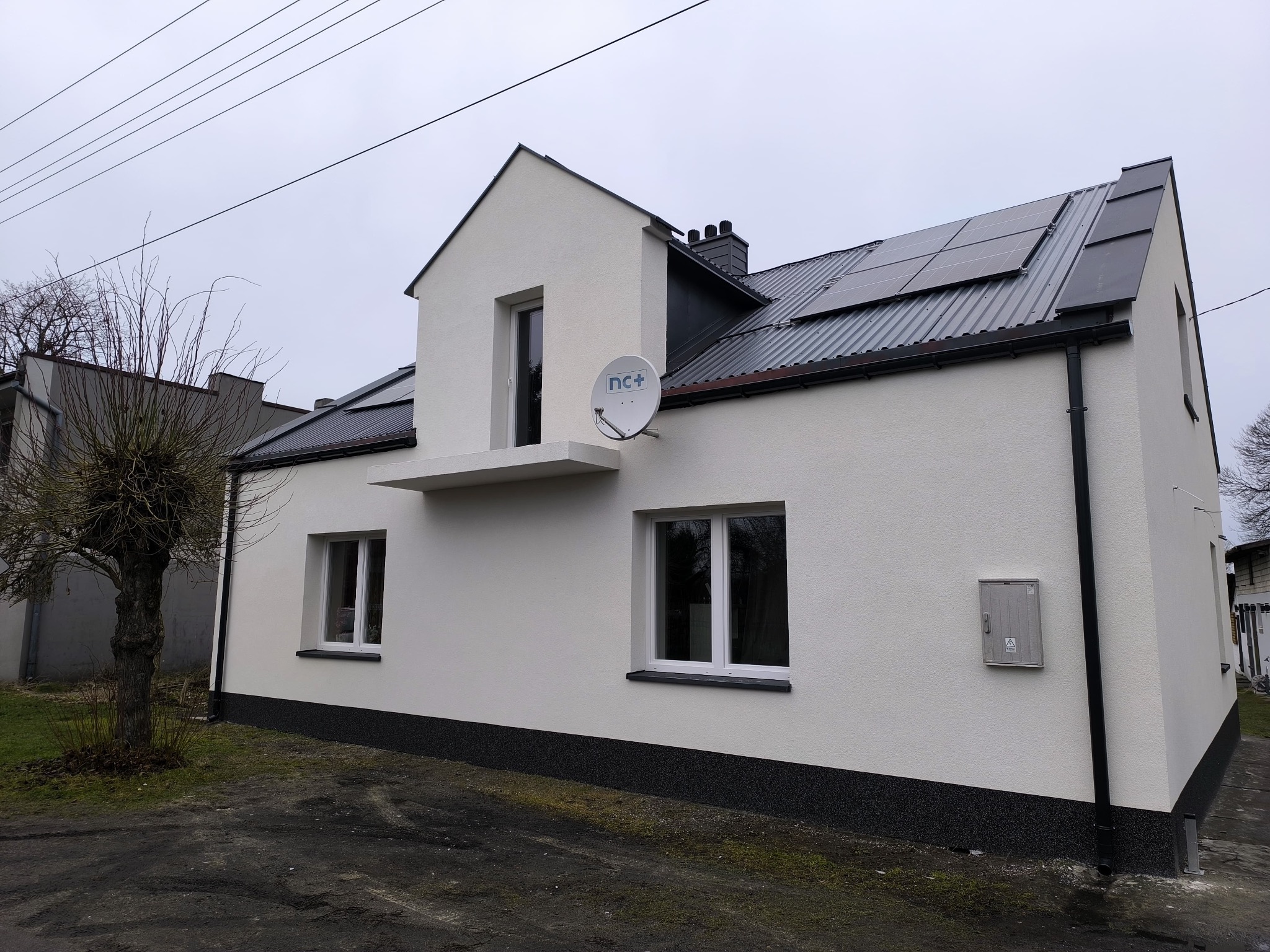 Biały dom z panelami słonecznymi na dachu i anteną satelitarną. Nowa elewacja z czarnym cokołem. Widok z zewnątrz w pochmurny dzień.