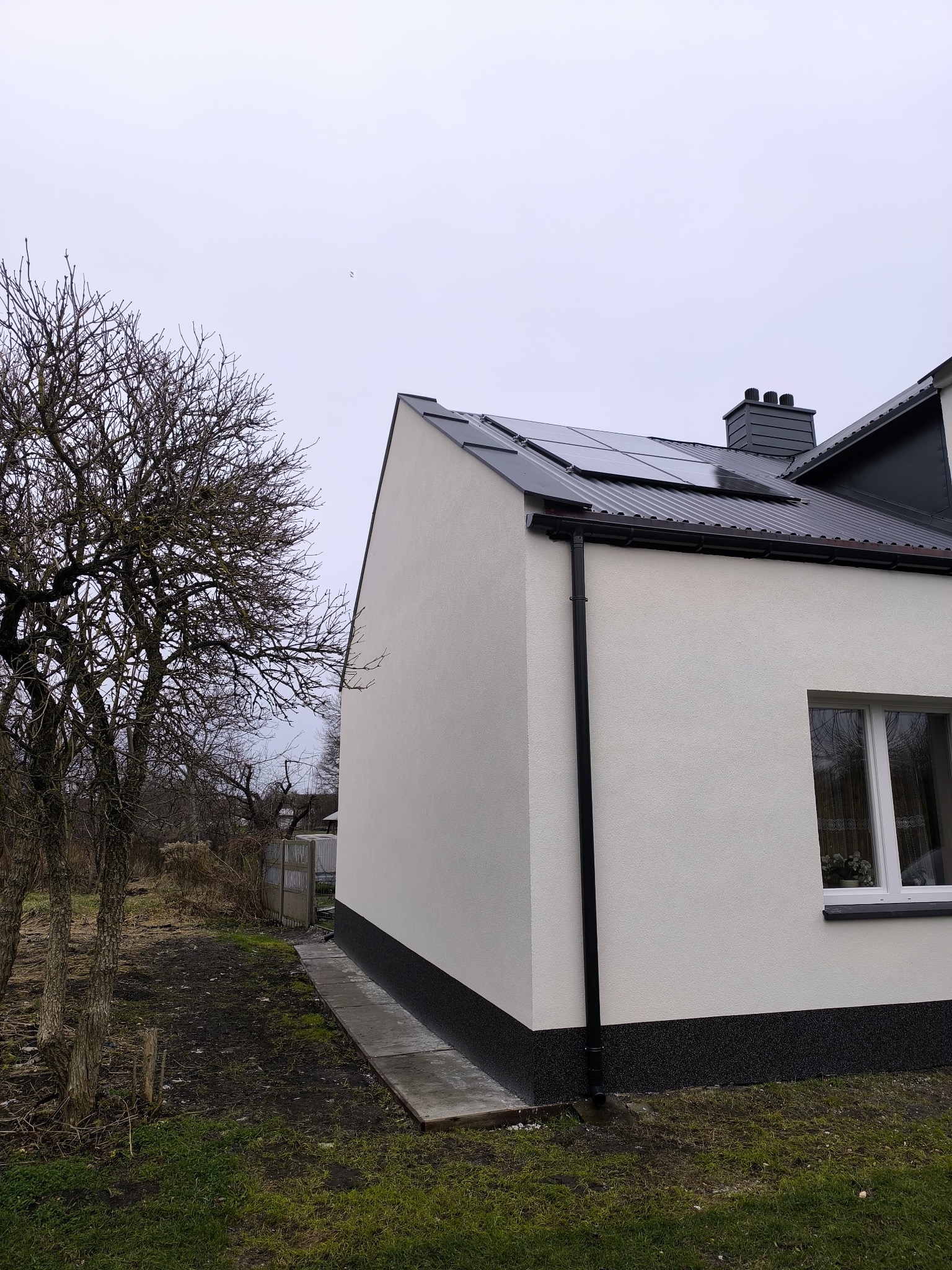 Ściana domu po tynkowaniu z widoczną elewacją w kolorze białym, czarną podmurówką i panelami słonecznymi na dachu. Obok drzewo bez liści i fragment ogrodu.