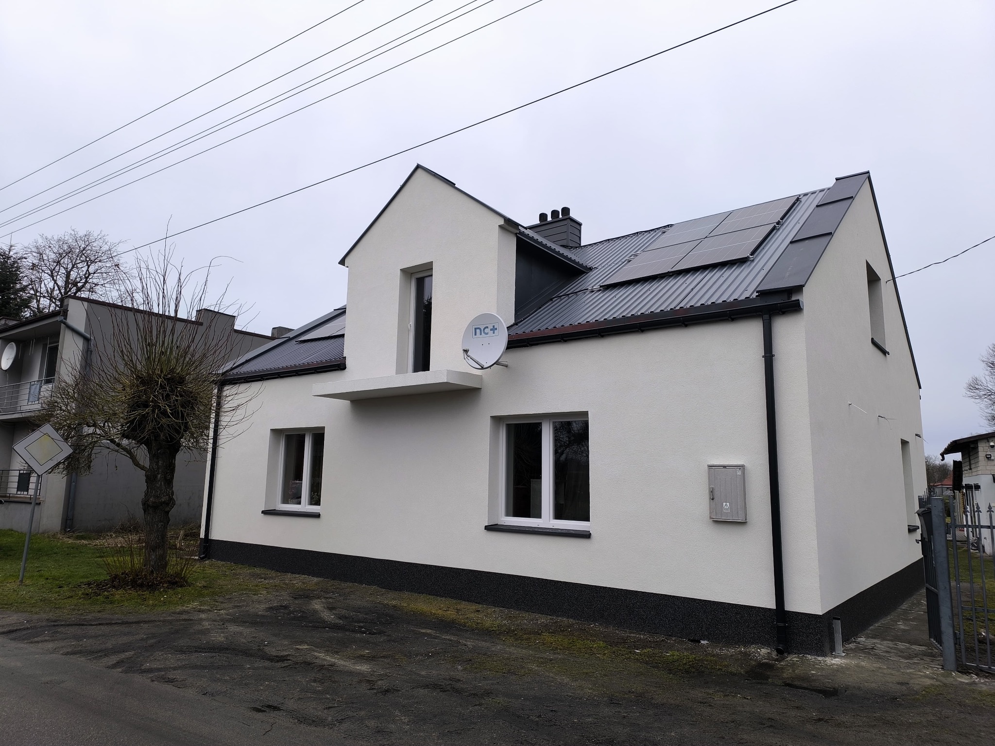 Biały dom jednorodzinny po remoncie elewacji w Zduńskiej Woli, z nową instalacją fotowoltaiczną na dachu i satelitą na ścianie. Widok z zewnątrz, pochmurny dzień.