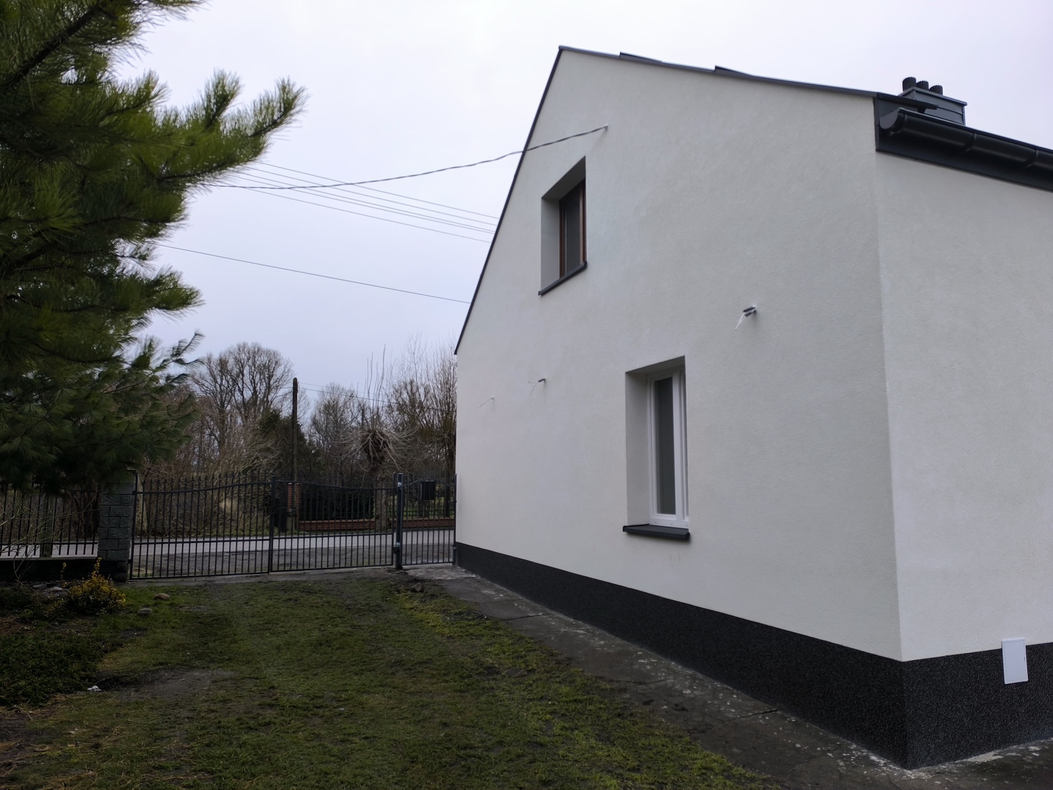 Biała elewacja domu z ciemną podmurówką, widok z boku na tle zieleni i metalowej bramy w pochmurny dzień. Widoczne dwa okna.