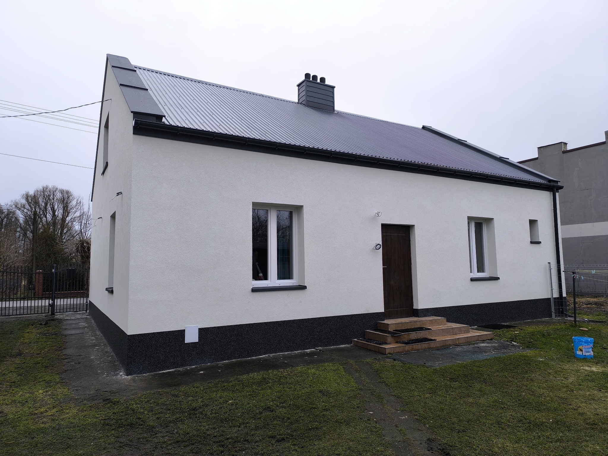 Biały dom jednorodzinny z szarą blachodachówką i ciemnym cokołem. Widoczne okna z białymi ramami i brązowe drzwi wejściowe. Dom po remoncie elewacji.