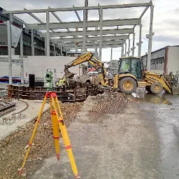 Ż&oacute;łty niwelator na tr&oacute;jnogu na pierwszym planie, w tle ż&oacute;łta koparka Komatsu i pracownicy przy budowie fundament&oacute;w pod konstrukcję stalową budynku.