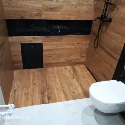 Nowoczesna łazienka z drewnopodobnymi płytkami na ścianach i podłodze, czarnym panelem dekoracyjnym z marmurkowym wzorem oraz białą, wiszącą toaletą.