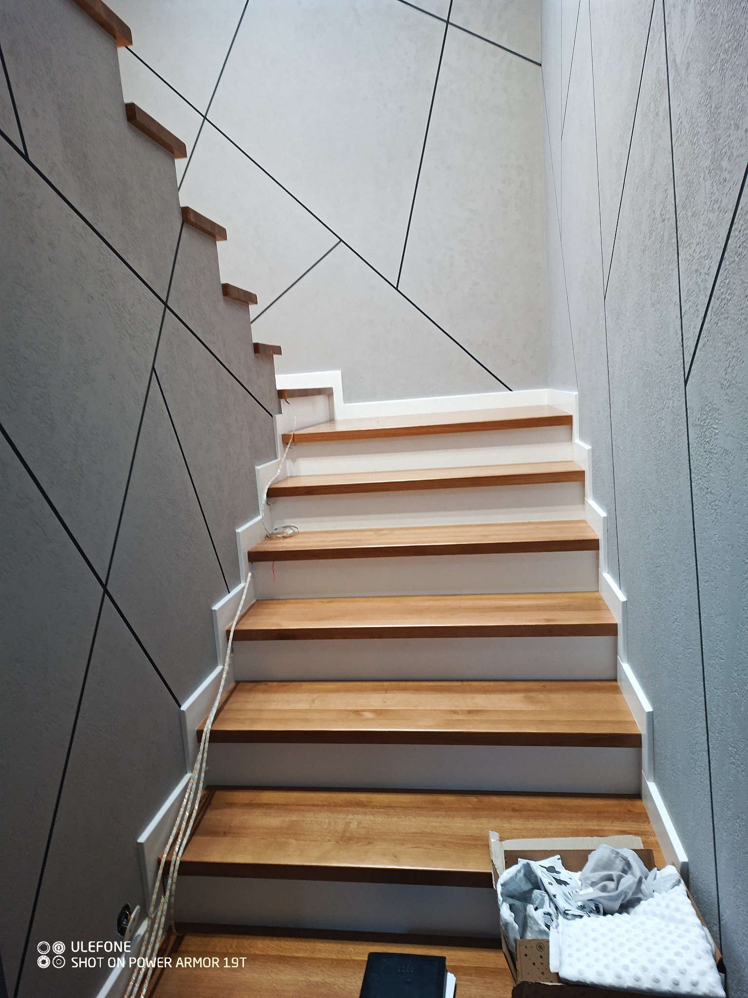 Drewniane schody z białymi podstopnicami, ściany z geometrycznym wzorem w odcieniach szarości, widoczna listwa ledowa i kartonowe pudło na dole schodów.