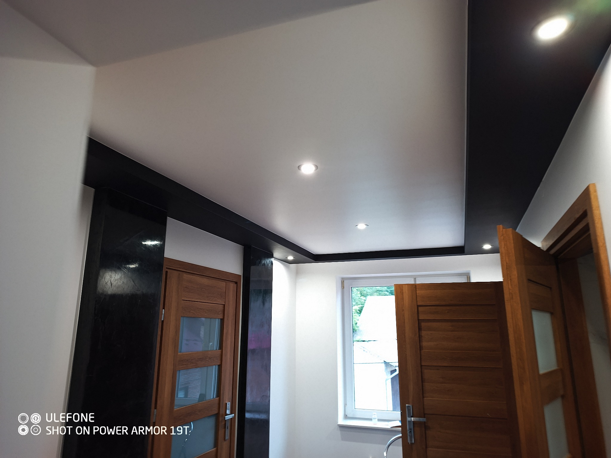 Biały sufit z czarnymi elementami dekoracyjnymi, oświetlenie punktowe, drewniane drzwi z przeszkleniami i białe ściany w nowocześnie wykończonym wnętrzu.