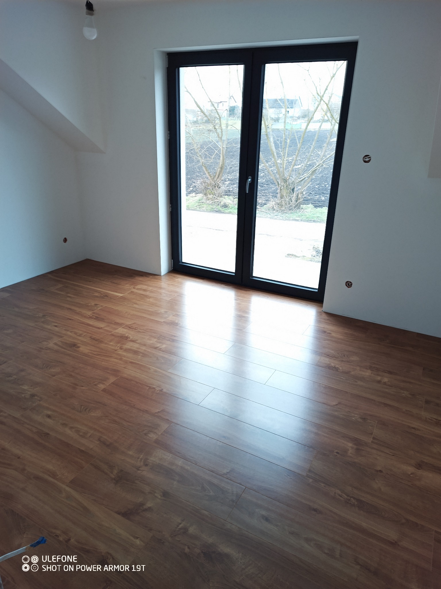 Nowo ułożony, błyszczący parkiet w pokoju z białymi ścianami i czarnymi drzwiami balkonowymi, widok na ogród za oknem.