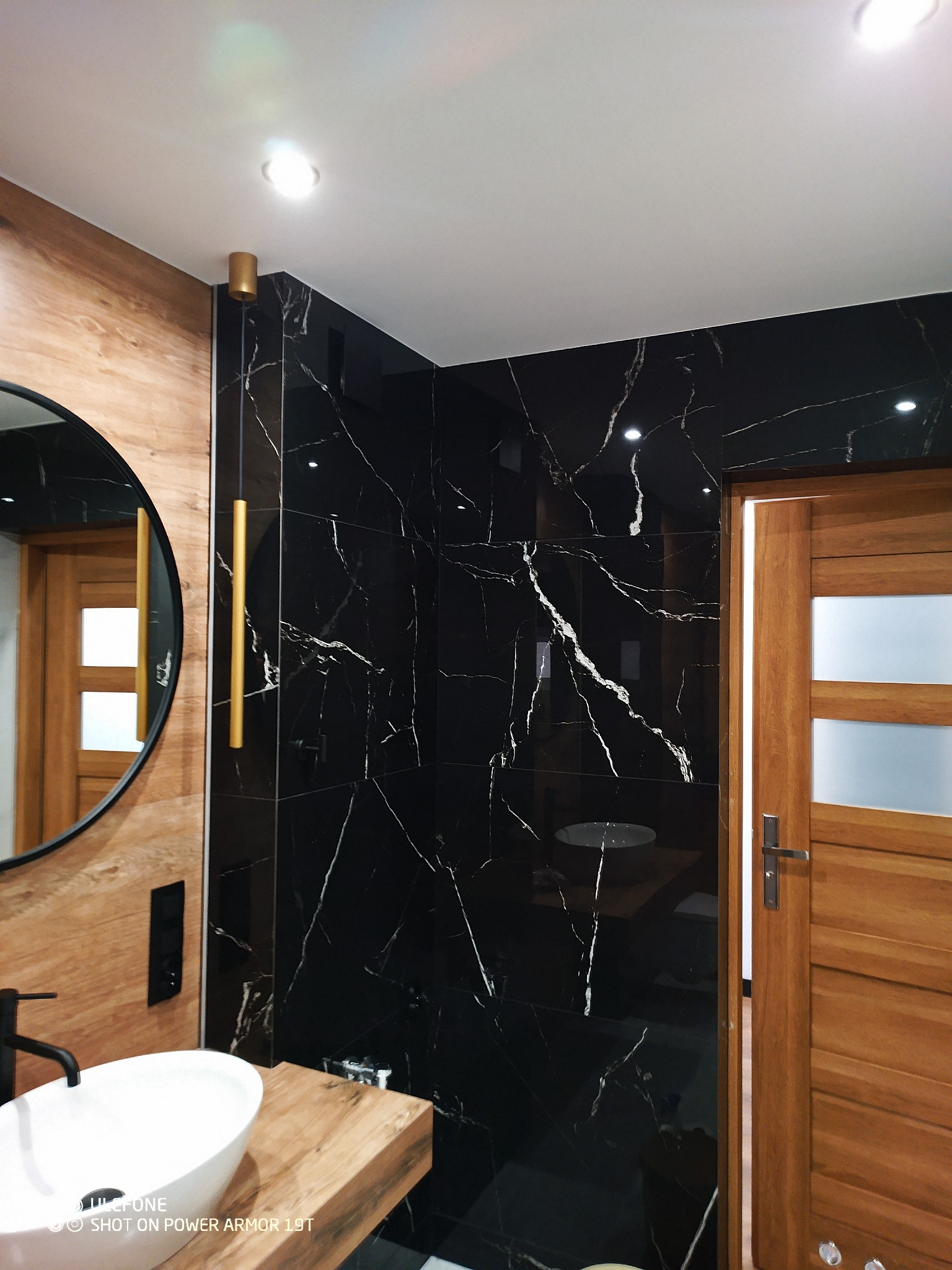 Nowoczesna łazienka z czarnymi płytkami imitującymi marmur, drewnianą umywalką i okrągłym lustrem z czarną ramą, oświetlona punktowymi lampami sufitowymi i złotą lampą wiszącą.