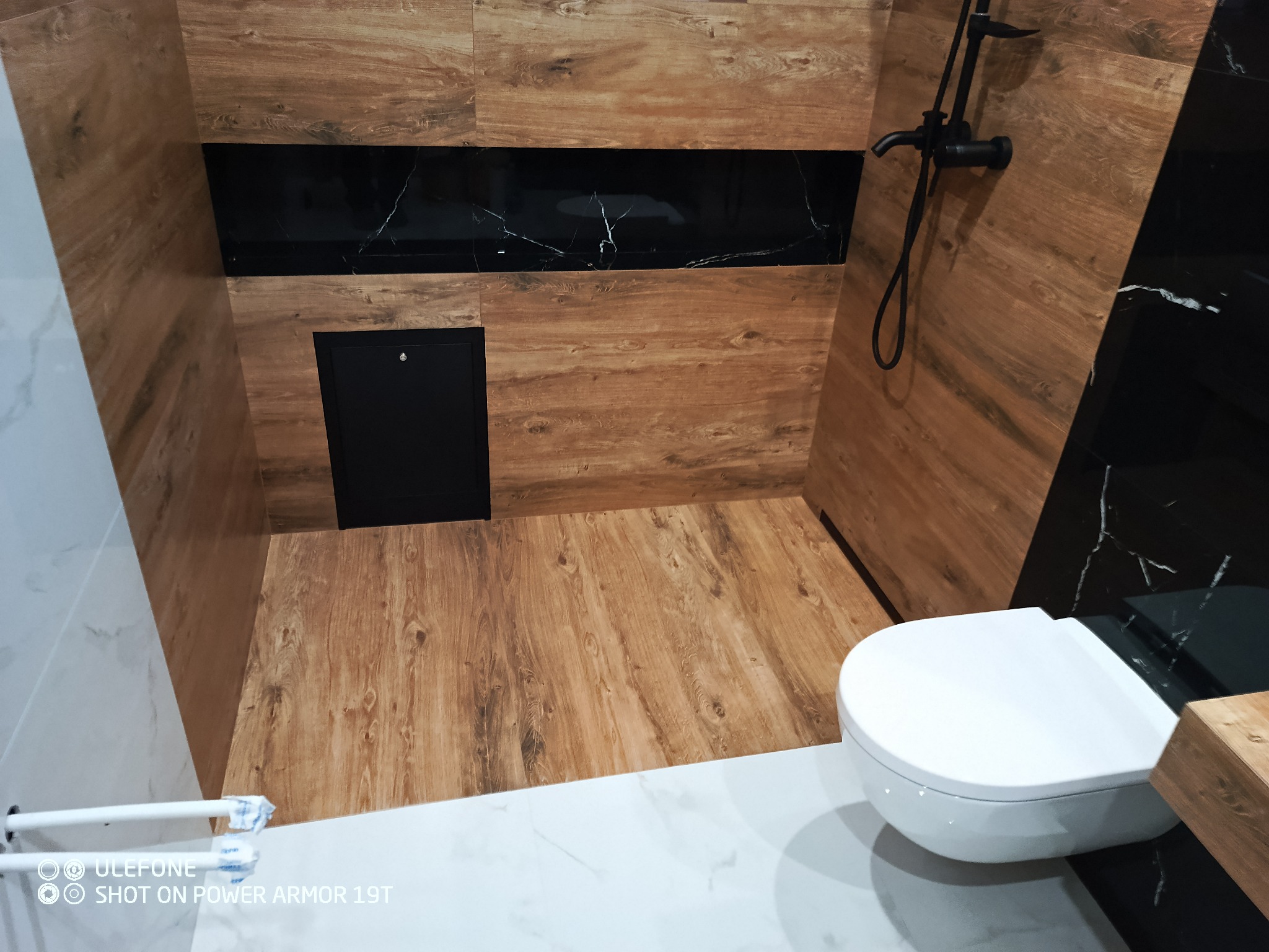 Nowoczesna łazienka z drewnopodobnymi płytkami na ścianach i podłodze, czarnym panelem dekoracyjnym z marmurkowym wzorem oraz białą, wiszącą toaletą.