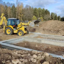 Żółta koparko-ładowarka New Holland przy wykopie pod fundamenty z wylanym betonem, widoczne drewniane paliki pomiarowe, w tle drzewa i błękitne niebo.