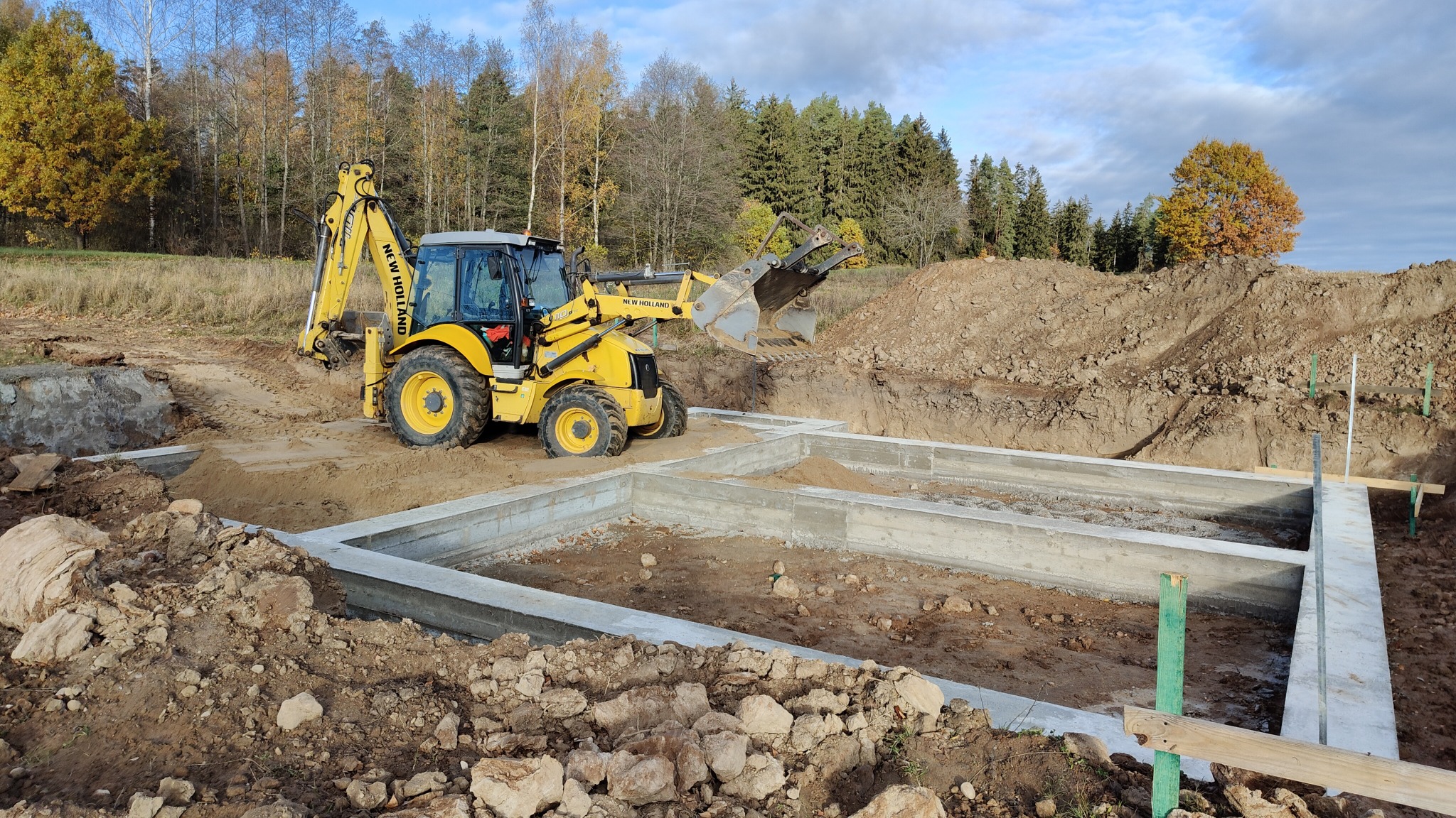 Żółta koparko-ładowarka New Holland przy wykopie pod fundamenty z wylanym betonem, widoczne drewniane paliki pomiarowe, w tle drzewa i błękitne niebo.