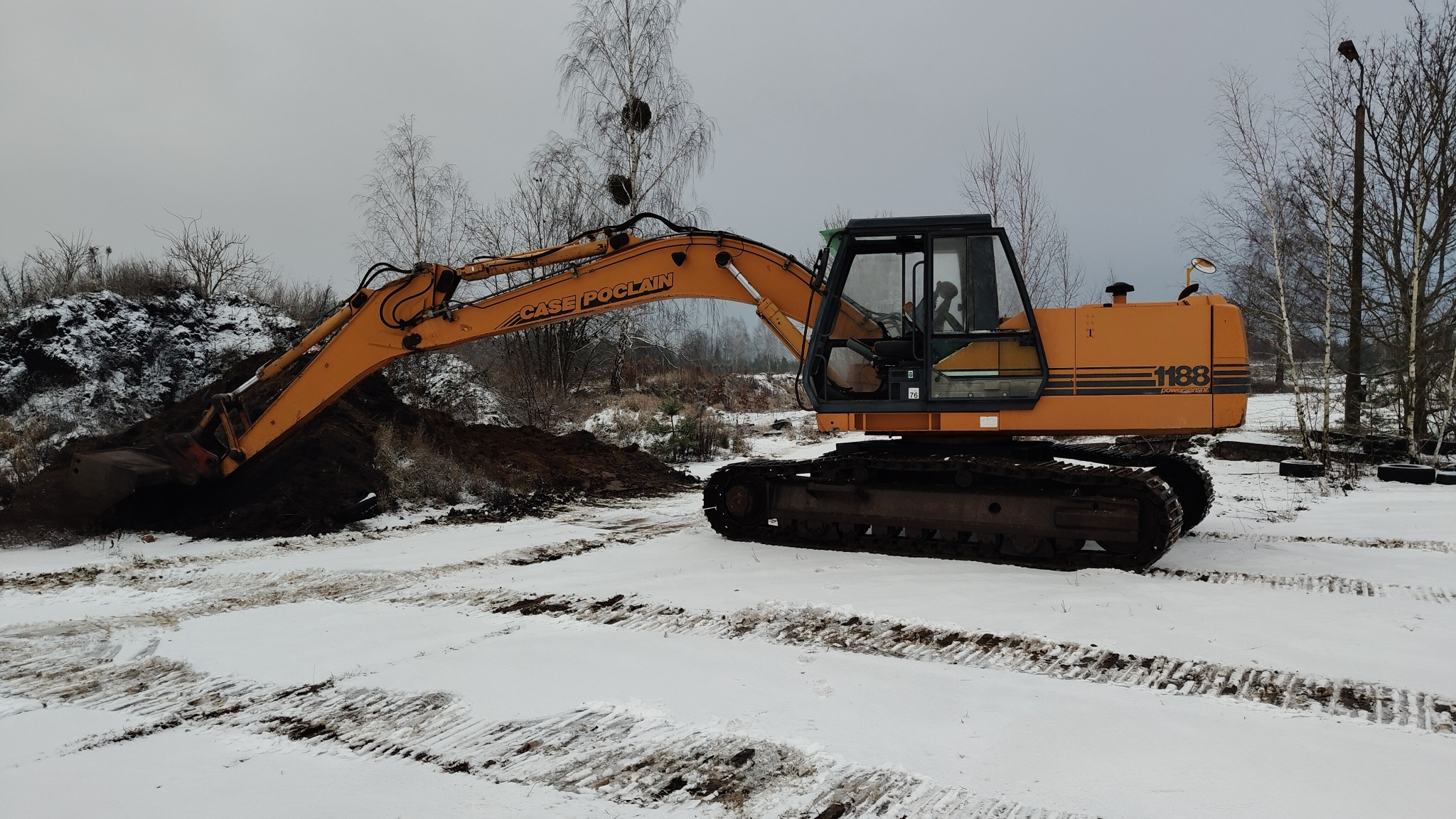 Pomarańczowa koparka Case Poclain 1188 nabiera ziemię z pryzmy na zaśnieżonym terenie, widoczne ślady gąsienic na śniegu.