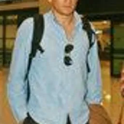 Mężczyzna w błękitnej koszuli, czapce z daszkiem i okularach przeciwsłonecznych zwisających z koszuli, z plecakiem, stojący na tle tablicy informacyjnej na lotnisku.