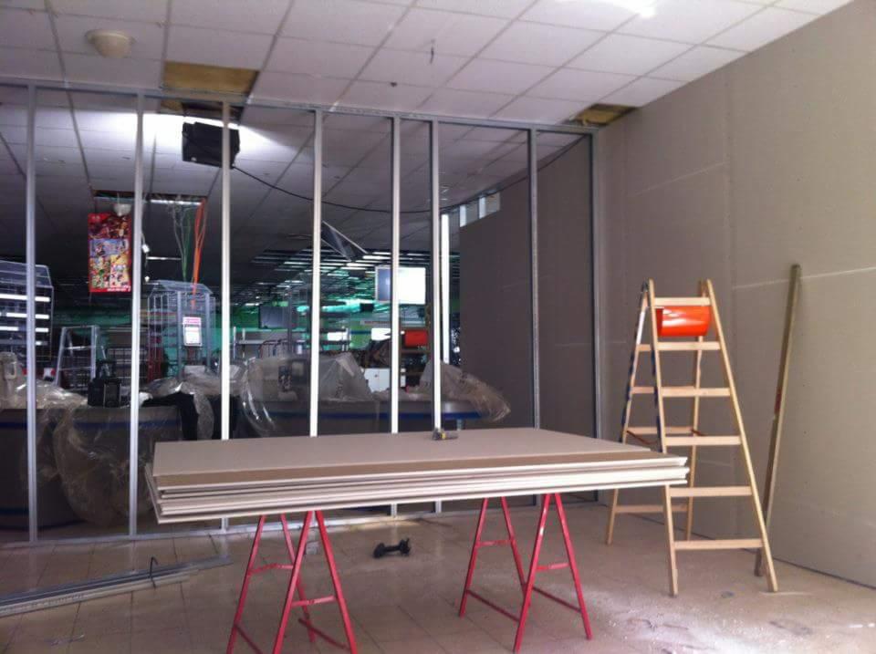 Wnętrze budynku w trakcie remontu: metalowa konstrukcja ścian działowych, stos płyt gipsowo-kartonowych na czerwonych kozłach, drabina drewniana z pomarańczowym wiadrem, widoczne elementy...