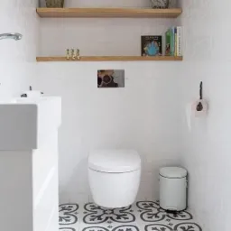 Biała łazienka z toaletą, umywalką, drewnianymi półkami z dekoracjami, białymi cegiełkami na ścianach i wzorzystymi płytkami podłogowymi w stylu retro.