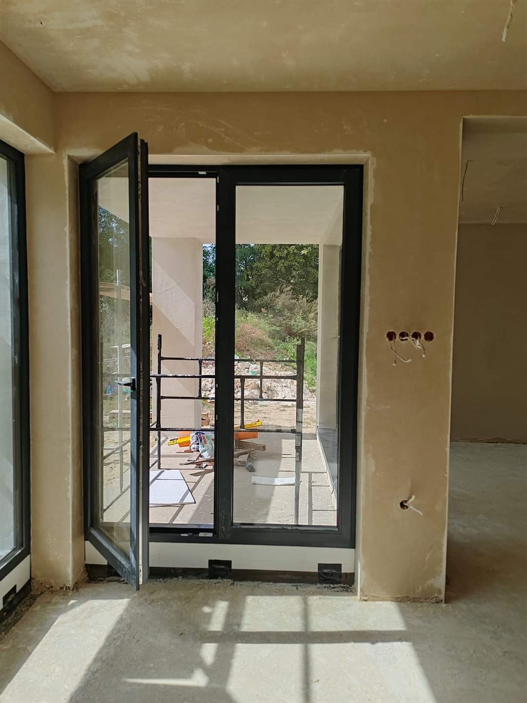 Wnętrze w trakcie prac wykończeniowych: surowe tynki na ścianach, otwarte okno balkonowe z czarną ramą, przez które widać rusztowanie i zieleń.