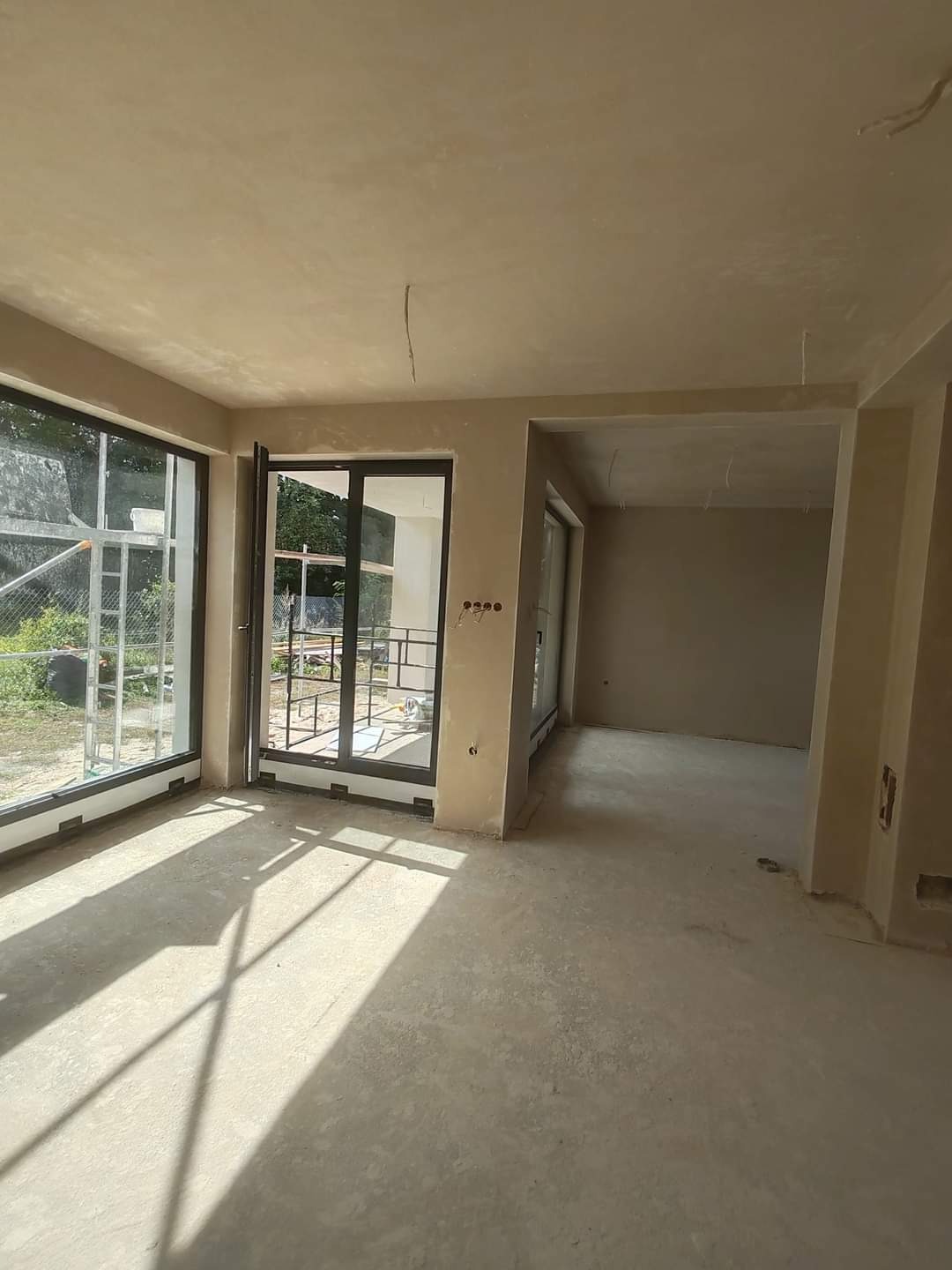 Wnętrze surowego pomieszczenia z dużymi oknami i świeżo nałożonymi tynkami na ścianach i suficie, widoczne wystające kable elektryczne.