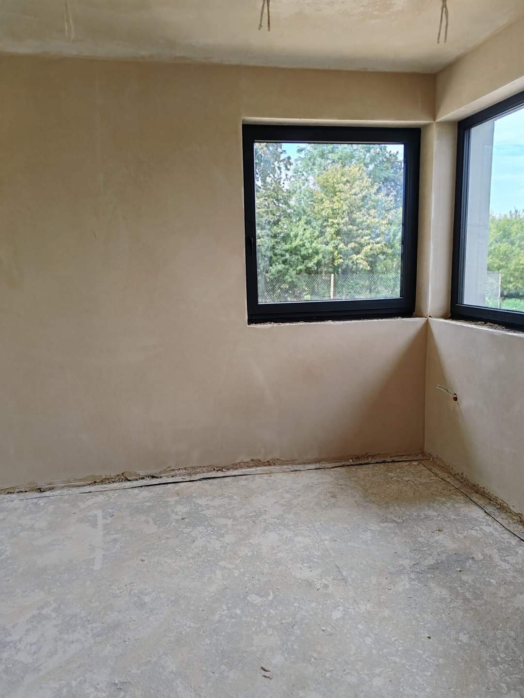 Surowe wnętrze z oknami w czarnych ramach i świeżo nałożonym tynkiem na ścianach, widoczne niedoskonałości na podłodze i wystające kable.