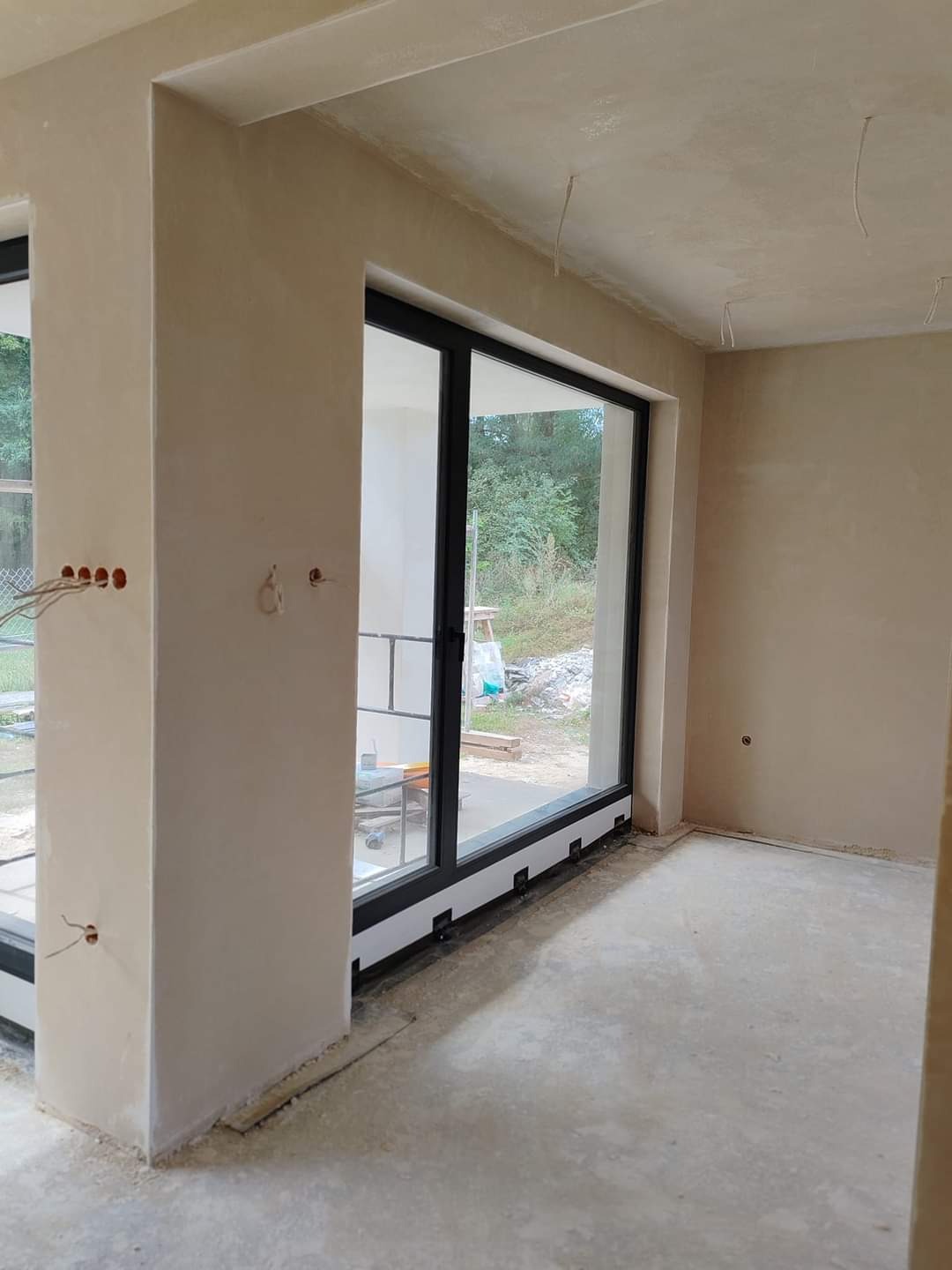 Wnętrze w trakcie tynkowania maszynowego: surowe ściany z widocznymi puszkami elektrycznymi, duże okno przesuwne z czarną ramą, betonowa podłoga z resztkami zaprawy.