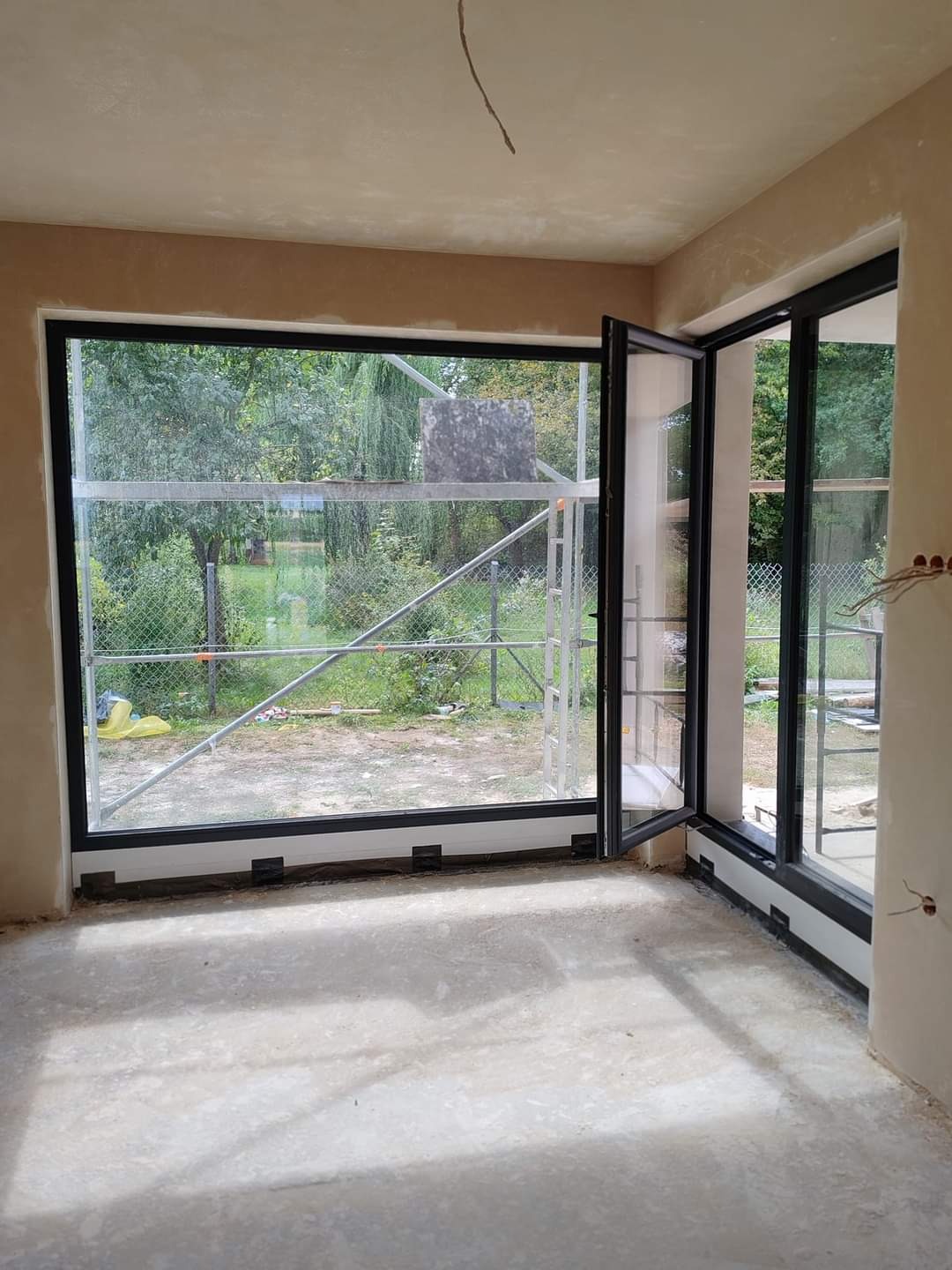 Wnętrze surowego pomieszczenia z dużymi oknami z czarnymi ramami, widok na zewnątrz na rusztowanie i zieleń, ściany w trakcie prac tynkarskich.