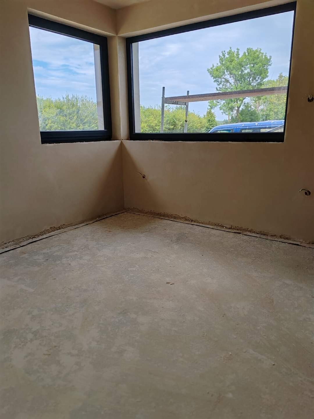Surowe wnętrze z dwoma oknami z czarnymi ramami, świeżo nałożone tynki na ścianach, widoczne wystające przewody elektryczne, podłoga betonowa, na zewnątrz widok na zieleń i rusztowanie.
