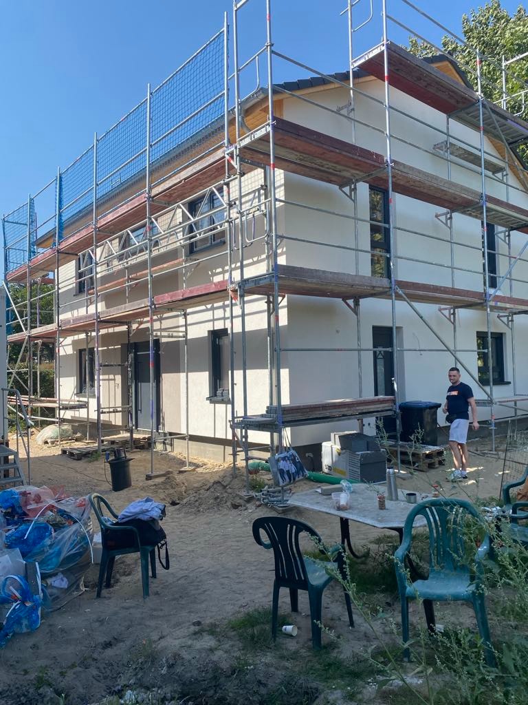 Dom w Dębnie w trakcie prac elewacyjnych, otoczony rusztowaniem i siatką ochronną, widoczna świeżo nałożona warstwa tynku, osoba w tle.