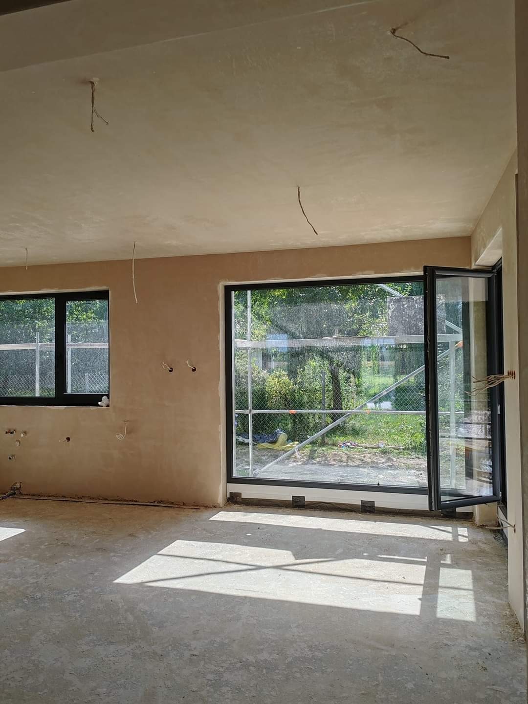 Surowe wnętrze z nałożonym tynkiem, widoczne czarne ramy okienne, otwarte drzwi balkonowe i wystające przewody elektryczne ze ścian i sufitu.