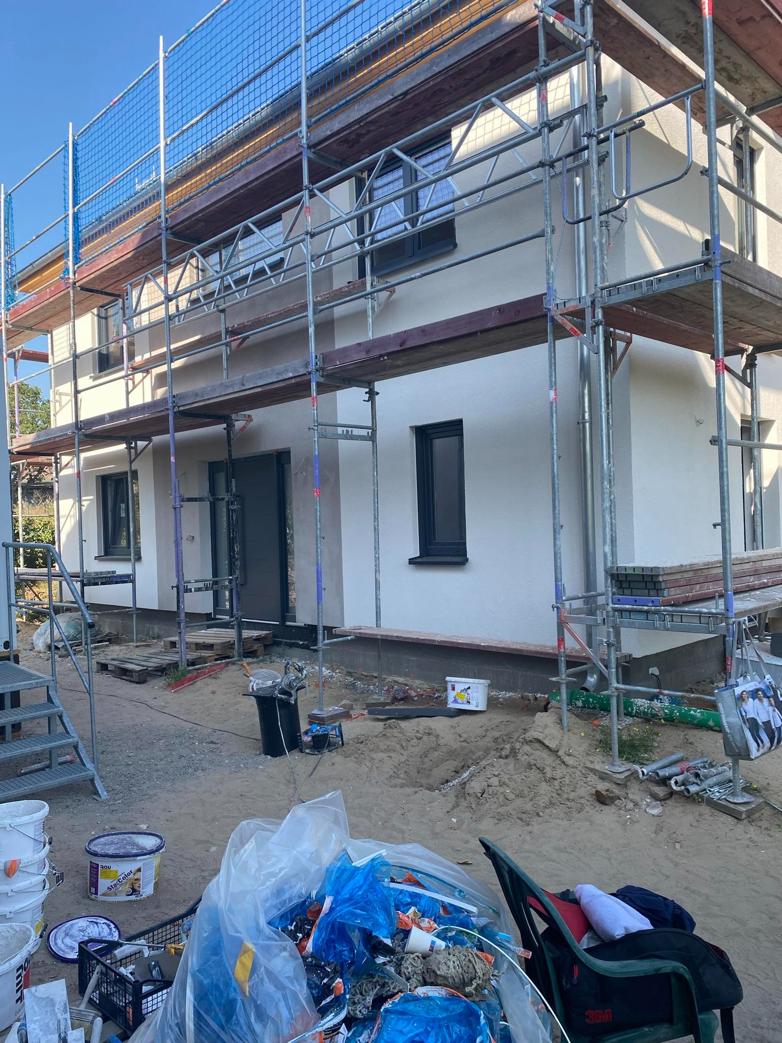 Dom jednorodzinny w trakcie prac elewacyjnych, widoczne rusztowania, świeżo położony tynk i przygotowane materiały budowlane na ziemi.