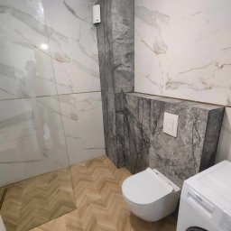 MED-BUD - Elegancka łazienka z marmurowymi płytkami i drewnianą podłogą w jodełkę. Wisząca toaleta i pralka Beko. Widoczny fragment kabiny prysznicowej.