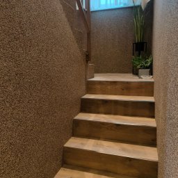 MED-BUD - Schody z drewnopodobnymi panelami i ścianami pokrytymi dekoracyjnym tynkiem, prowadzące w górę, z roślinami w tle i oknem z delikatną firanką.