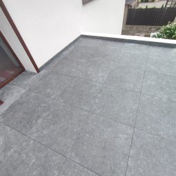 MED-BUD - Szare płytki na tarasie wentylowanym z wklejonymi cokołami, widok z góry, jasne ściany, okno. Detal wykończenia.