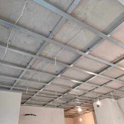 MED-BUD - Sufit podwieszany z metalową konstrukcją w trakcie montażu, widoczne kable i fragment ściany z instalacją. Surowy stan wykończenia wnętrza.