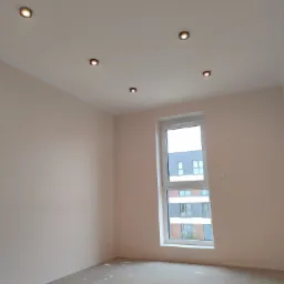 Pokój po nałożeniu gładzi, z widocznymi punktowymi lampami w suficie i oknem z widokiem na budynek.