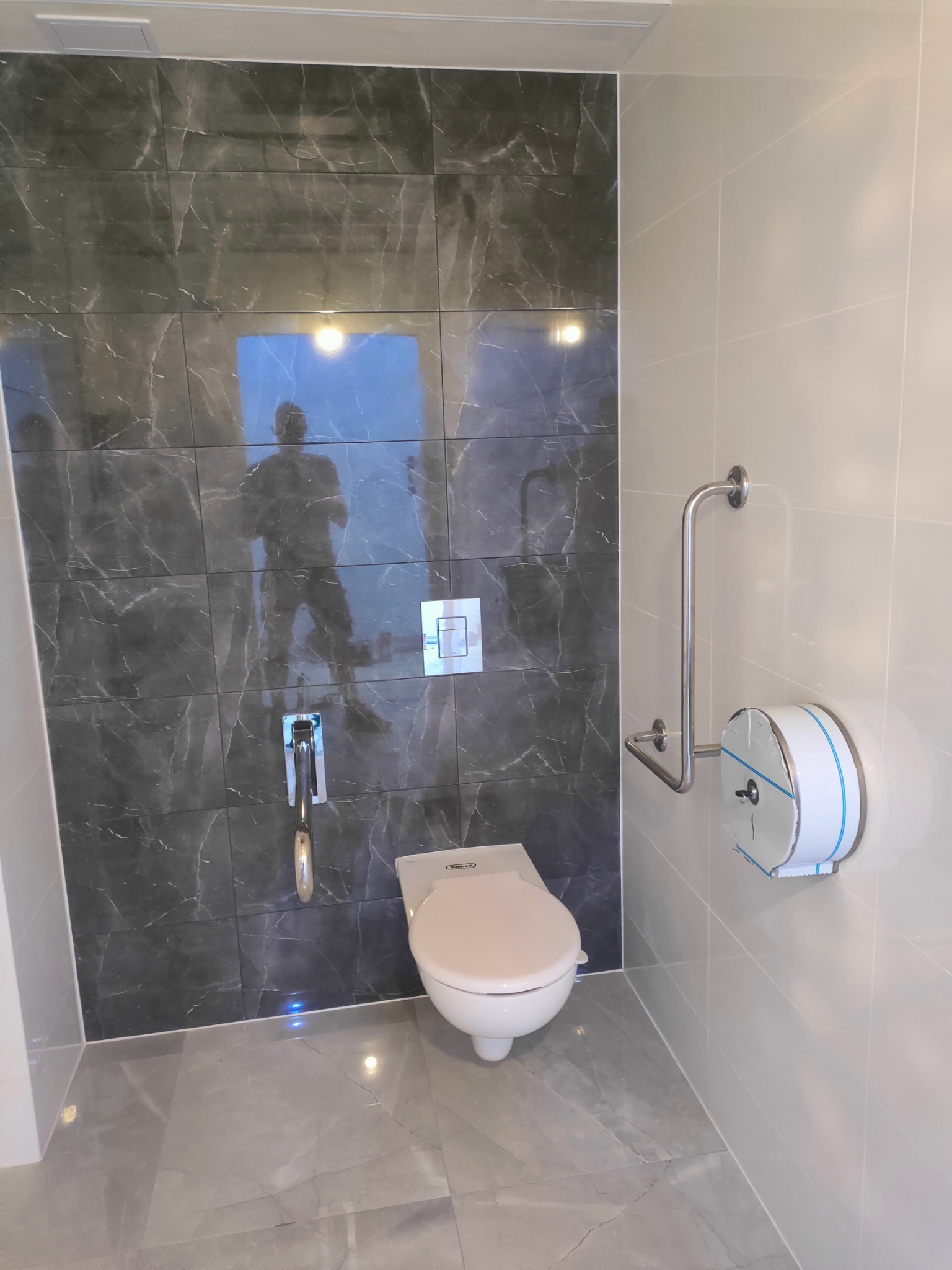 Nowoczesna łazienka z szarymi płytkami imitującymi marmur, toaleta z uchwytem dla niepełnosprawnych i dozownikiem papieru. Widoczne odbicie osoby w płytkach.