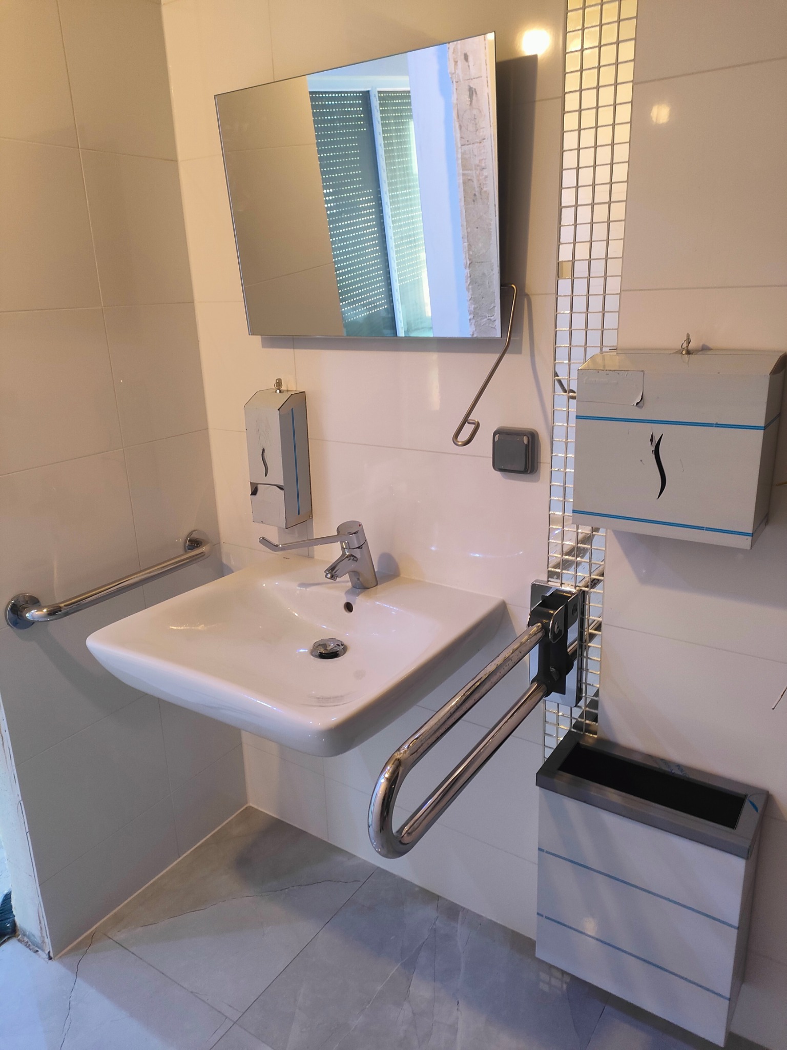 Funkcjonalna łazienka z umywalką i uchwytami dla niepełnosprawnych, jasne płytki, lustro i dozownik mydła, metalowe akcenty, nowoczesny design.