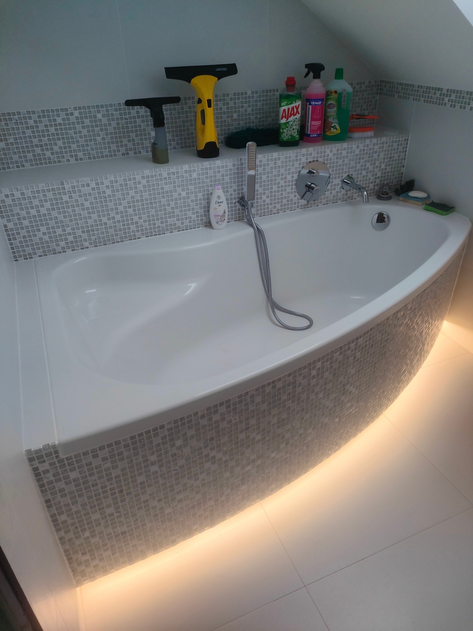 Nowoczesna, narożna wanna obudowana mozaiką z podświetleniem LED w łazience na poddaszu. Na półce nad wanną środki czystości i akcesoria.