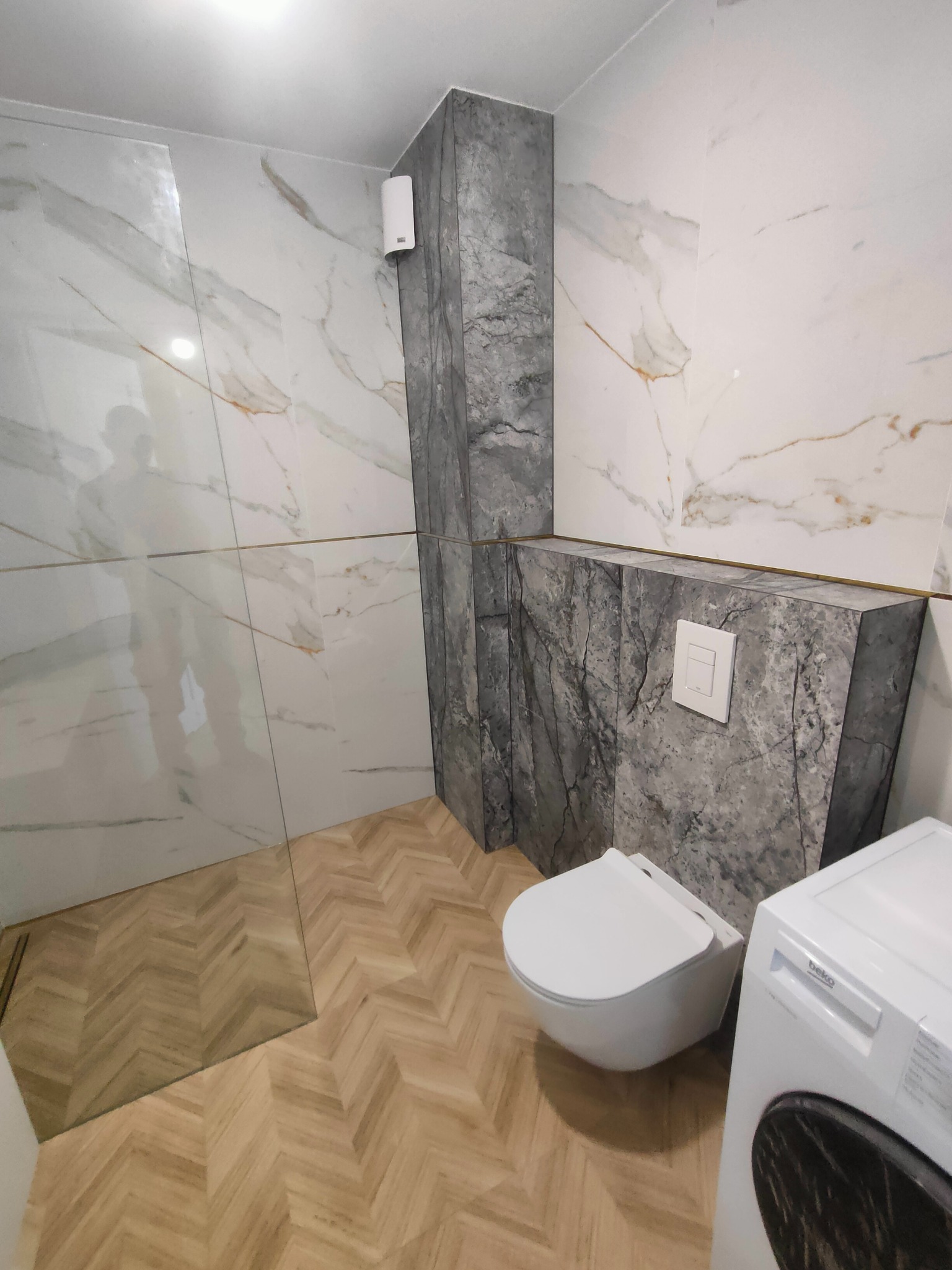 Elegancka łazienka z marmurowymi płytkami i drewnianą podłogą w jodełkę. Wisząca toaleta i pralka Beko. Widoczny fragment kabiny prysznicowej.