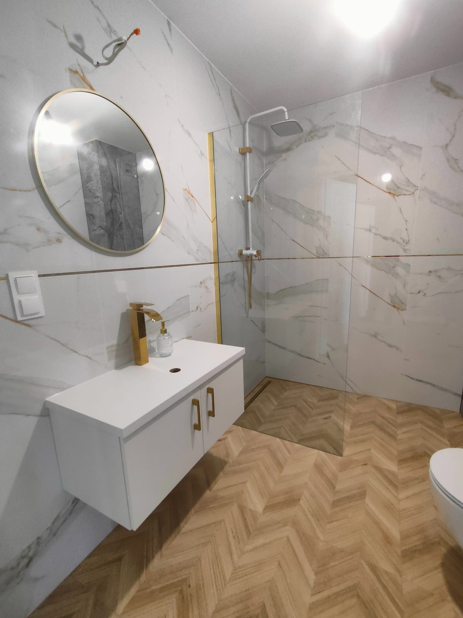 Elegancka łazienka po remoncie: białe marmurowe płytki z złotymi akcentami, armatura w kolorze złota, wzór jodełki na podłodze, kabina prysznicowa ze szkła.