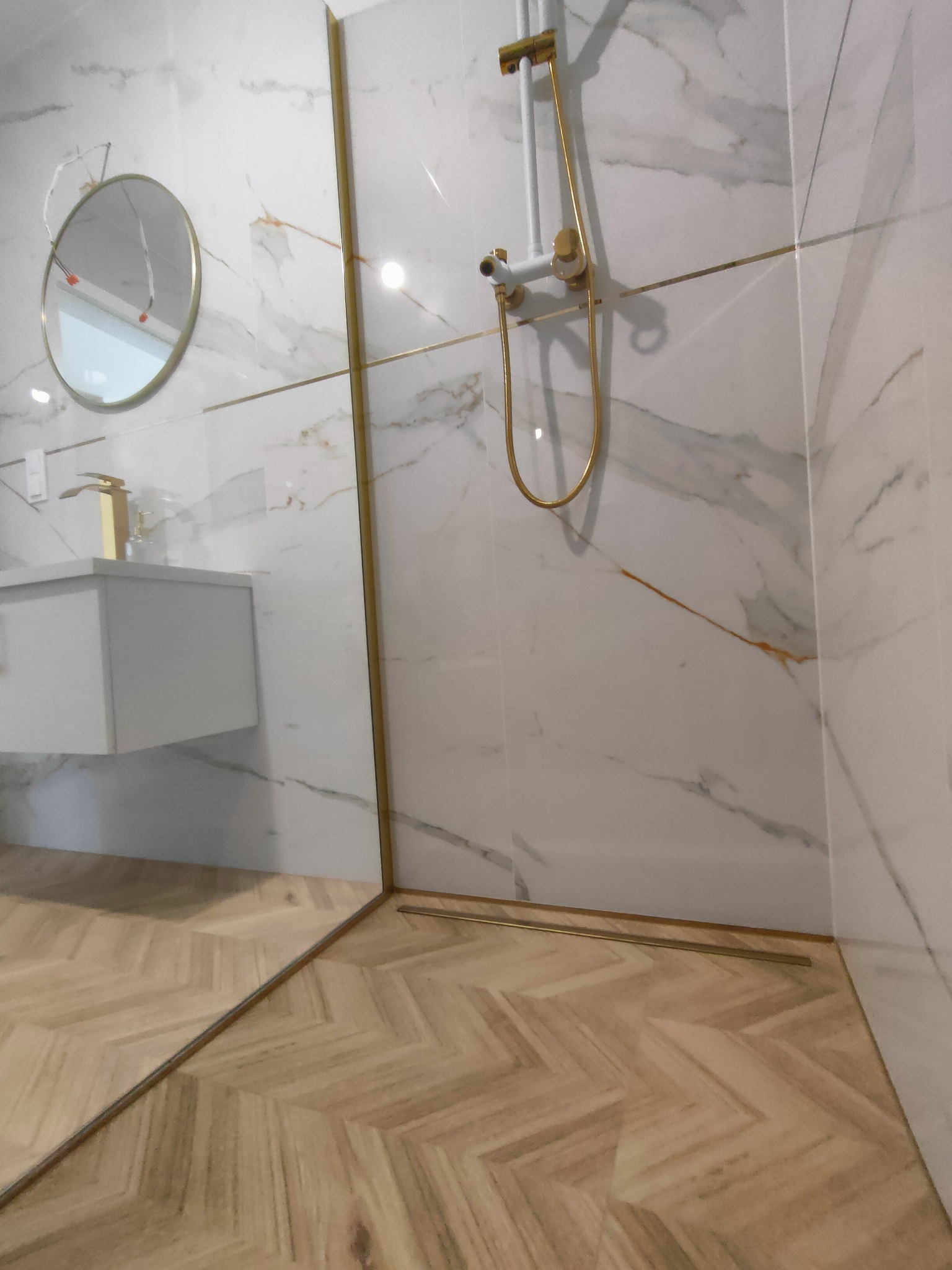 Wykończona łazienka z marmurowymi płytkami, złotymi akcentami i drewnianą podłogą w jodełkę. Widoczna kabina prysznicowa i umywalka z okrągłym lustrem.