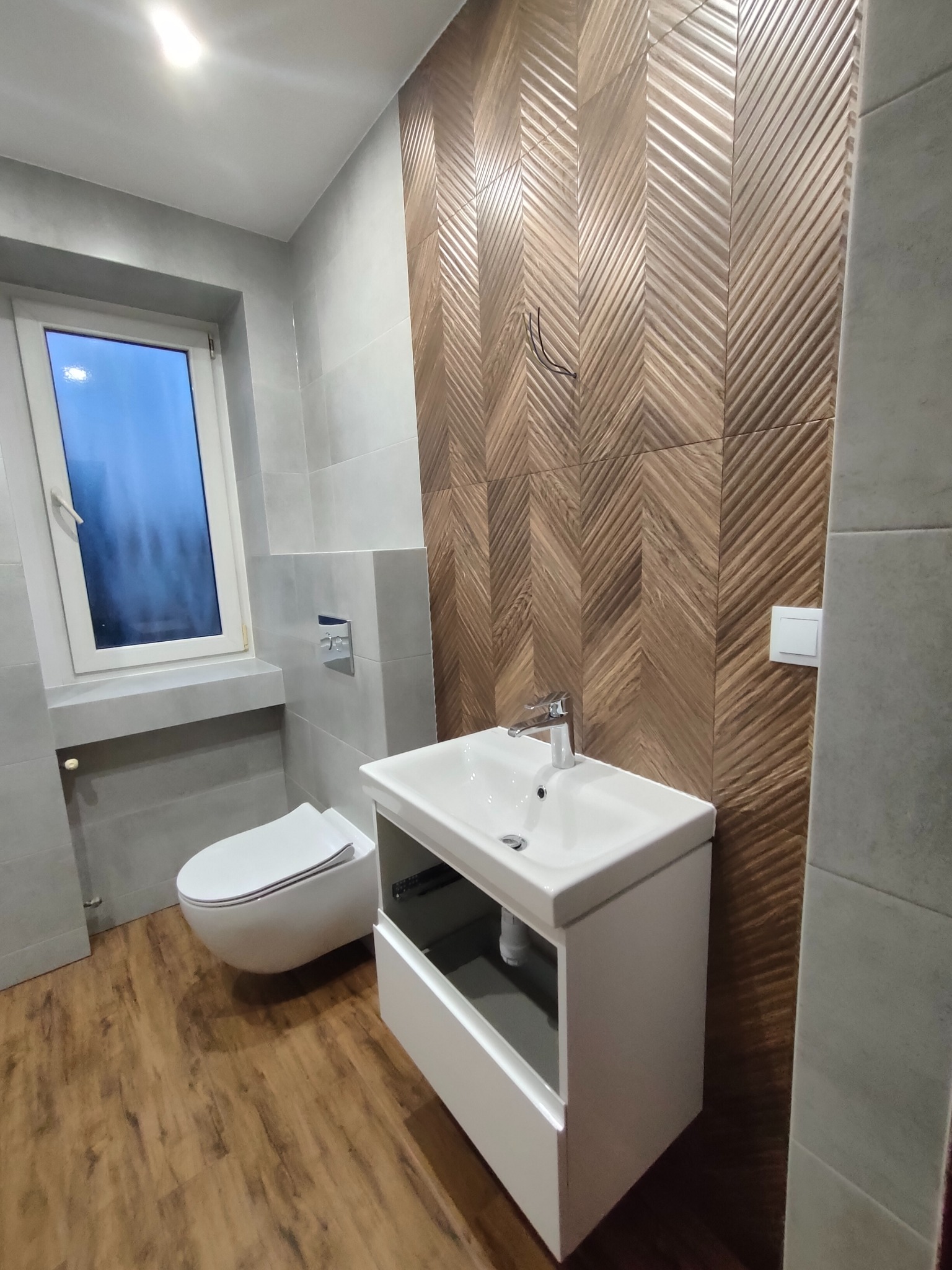 Nowoczesna łazienka po remoncie: drewnopodobna ściana w jodełkę, biała umywalka z szafką i podwieszana toaleta. Szare płytki i drewniana podłoga tworzą spójny design.