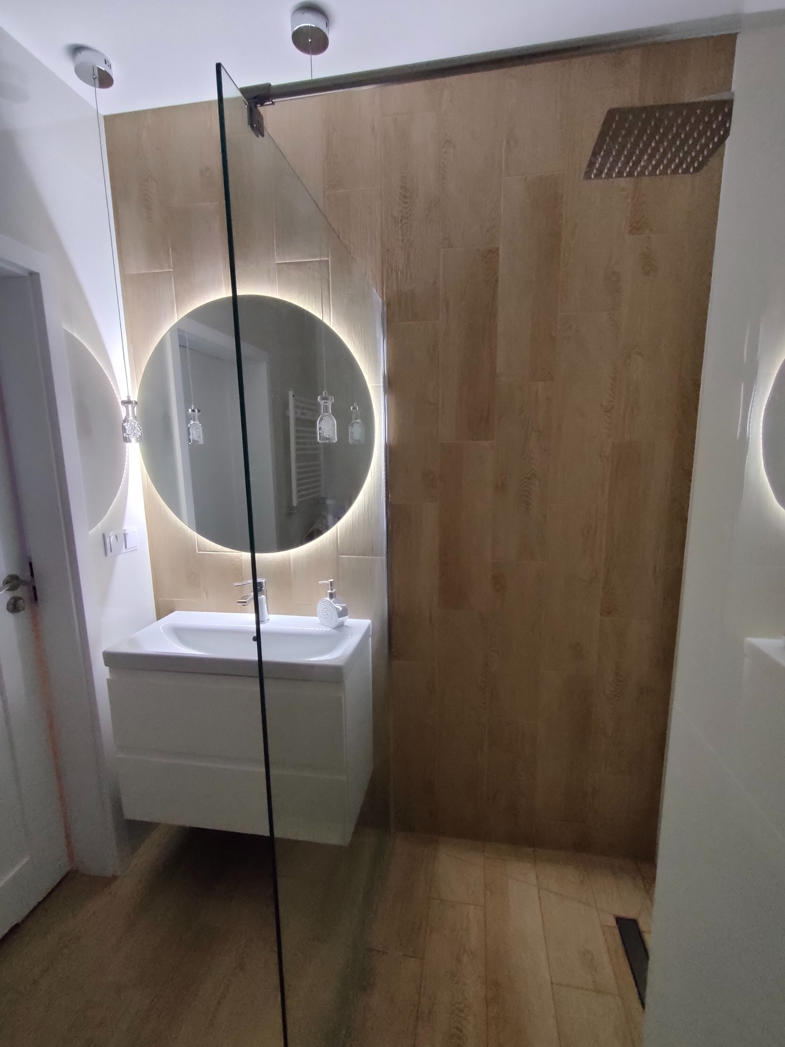 Nowoczesna łazienka po remoncie: drewnopodobne płytki, szklana kabina, umywalka z podświetlanym lustrem. Minimalistyczny design i eleganckie wykończenie.