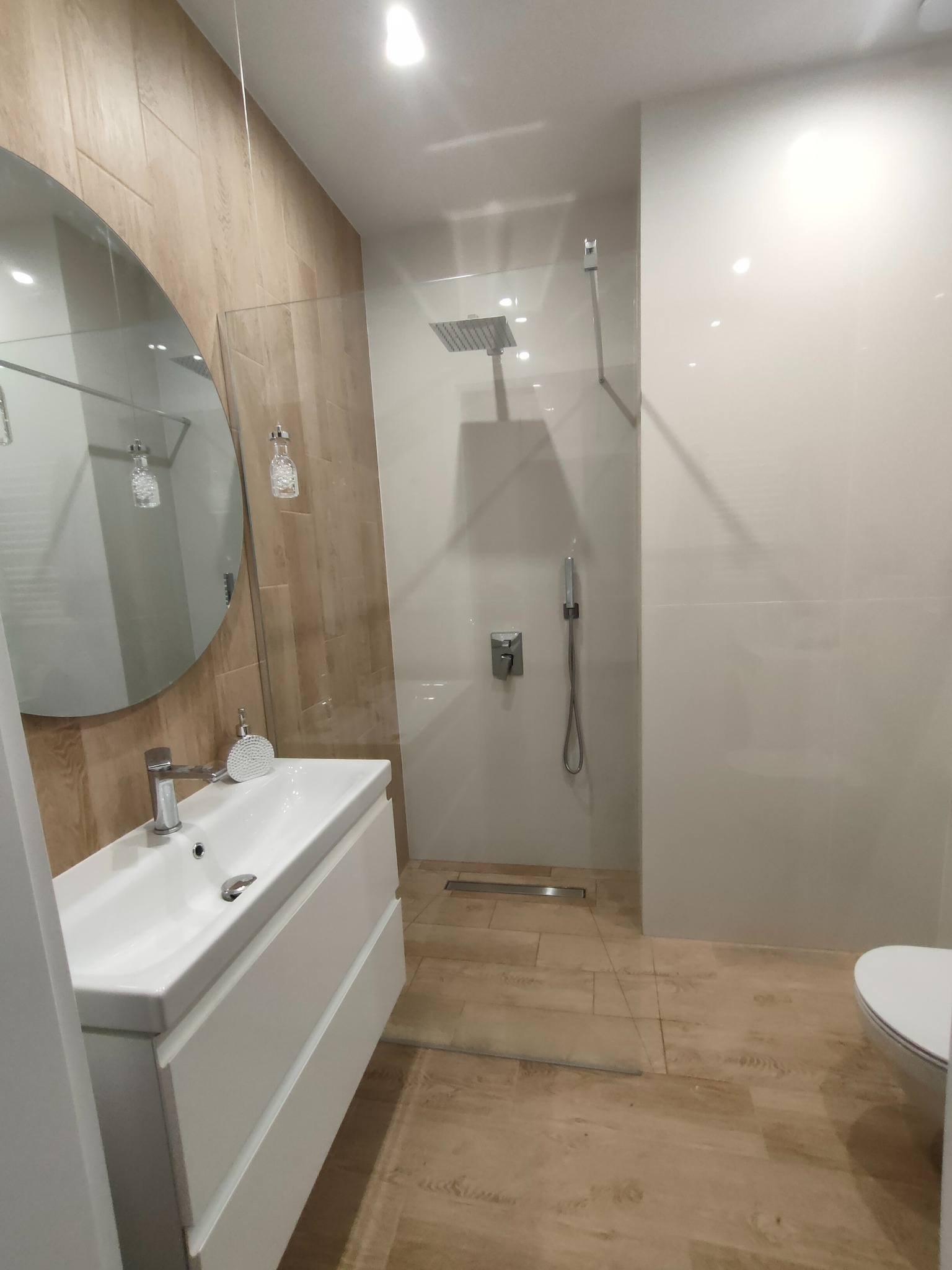 Nowoczesna łazienka po remoncie: kabina typu walk-in, okrągłe lustro nad białą szafką z umywalką, płytki imitujące drewno na podłodze i ścianie.