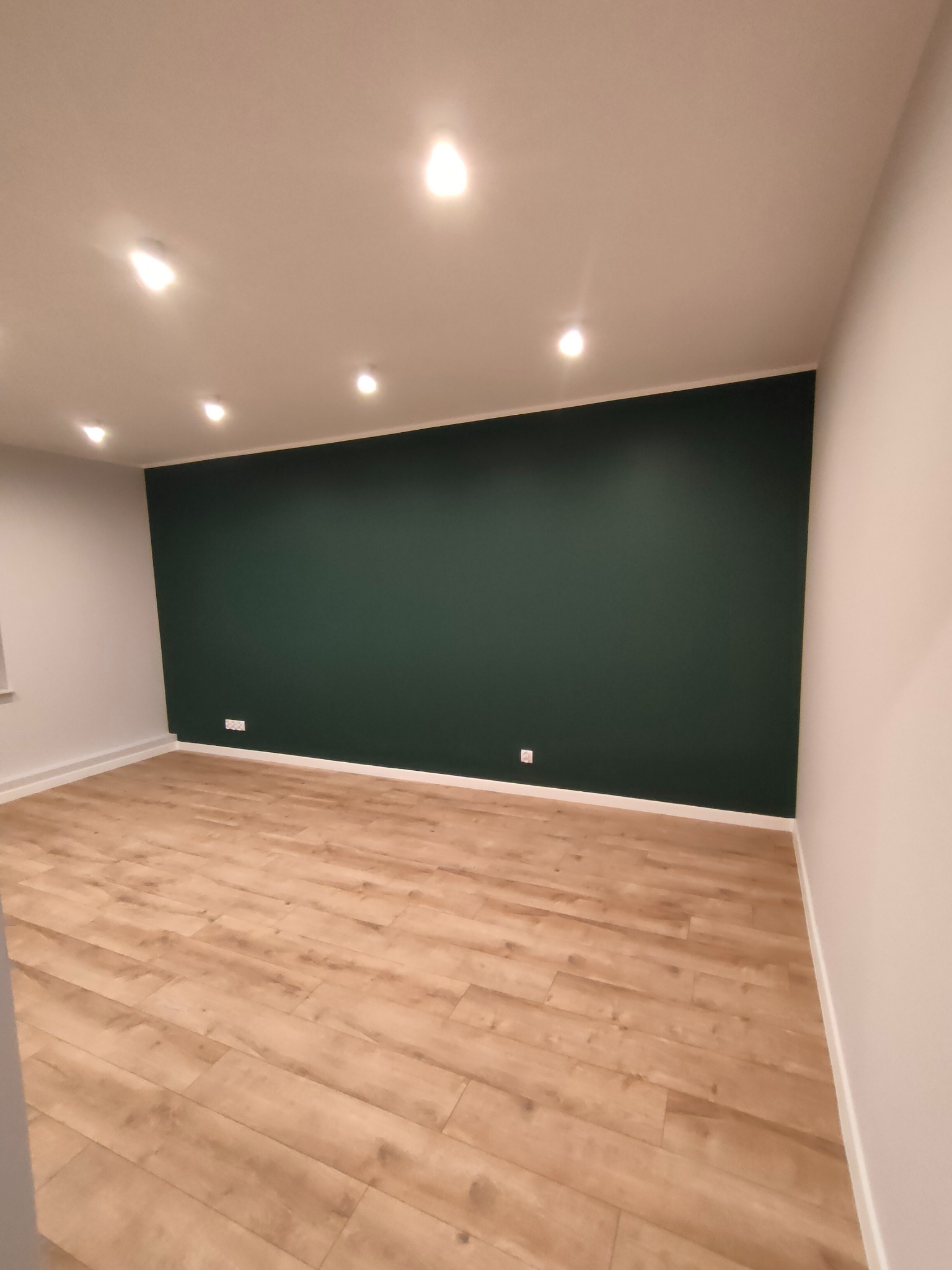 Pokój po remoncie: ściana pomalowana na butelkową zieleń, jasne panele podłogowe, sufit z oświetleniem punktowym. Widoczne gniazdka elektryczne przy podłodze.