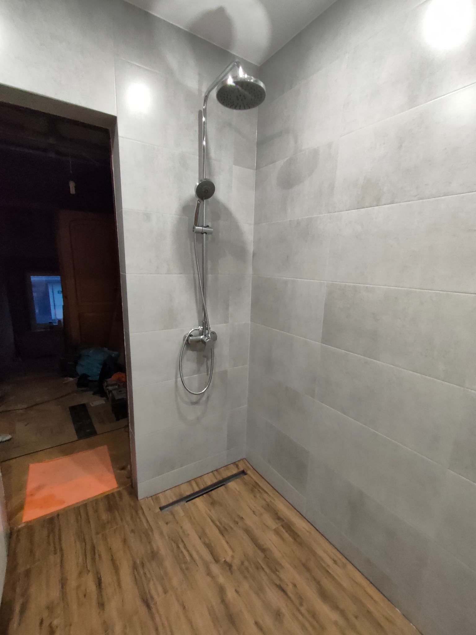 Wykończona łazienka z drewnopodobną podłogą i szarymi płytkami ściennymi. Widoczna kabina prysznicowa z deszczownicą. W tle widoczne niedokończone prace.