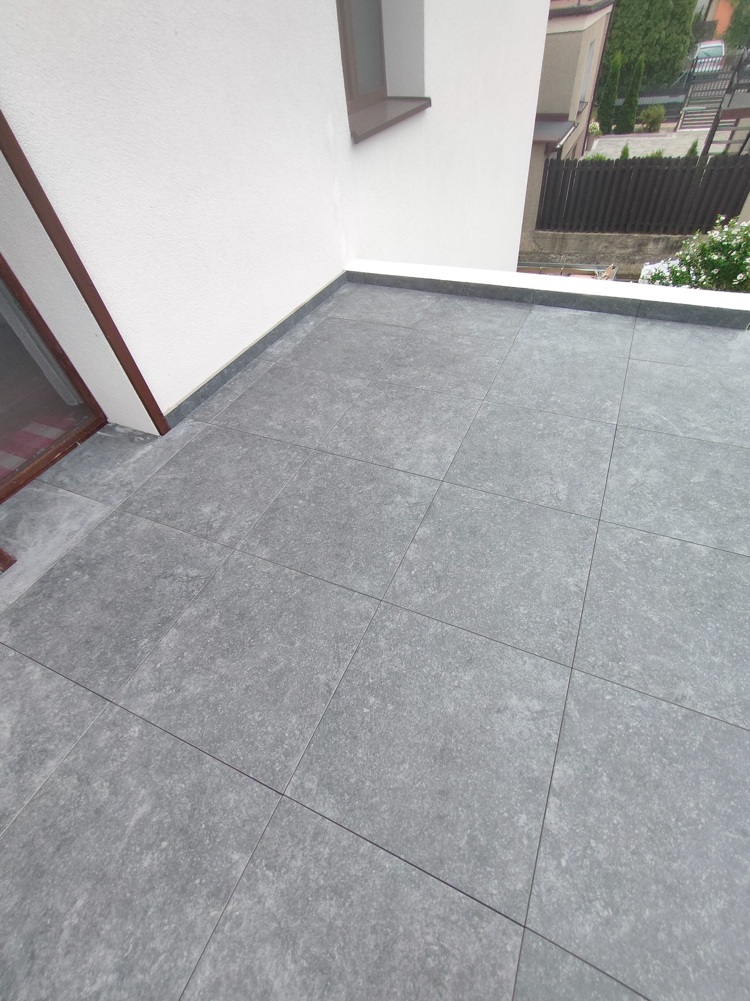 Szare płytki na tarasie wentylowanym z wklejonymi cokołami, widok z góry, jasne ściany, okno. Detal wykończenia.