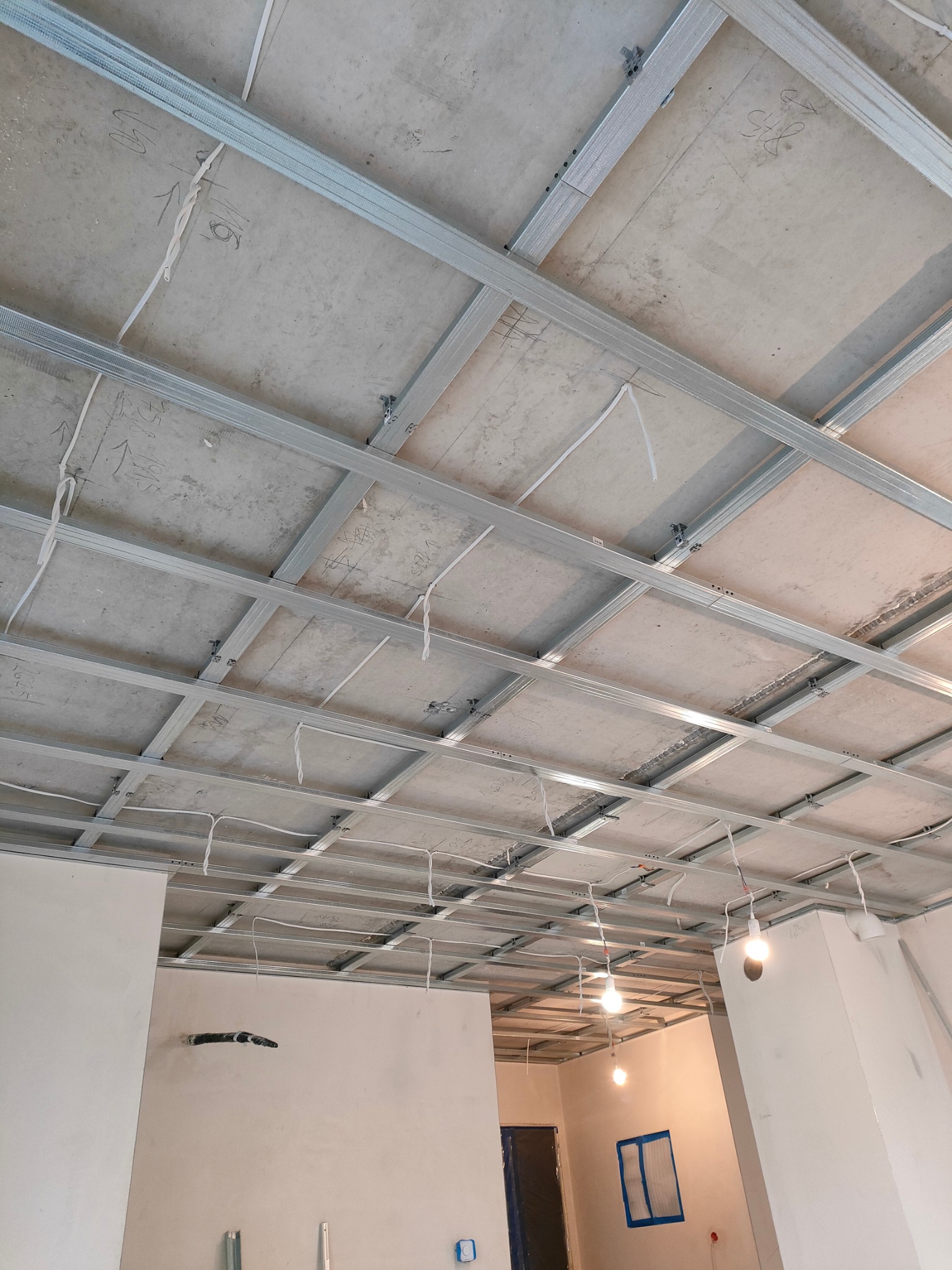 Sufit podwieszany z metalową konstrukcją w trakcie montażu, widoczne kable i fragment ściany z instalacją. Surowy stan wykończenia wnętrza.