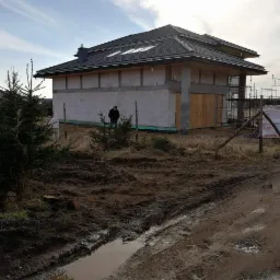 Dom w trakcie budowy z czarną dachówką na dachu, widoczne rusztowania i osoba stojąca obok, teren wokół domu błotnisty.