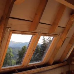 Dwa okna dachowe w drewnianych ramach z widokiem na zalesione wzgórza, widoczne elementy konstrukcji dachu i ocieplenia.