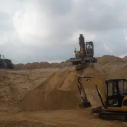 Żółta koparka CAT 308E CR w trakcie niwelacji terenu piaszczystego, w tle widoczny traktor z przyczepą wypełnioną piaskiem oraz druga koparka, pochmurne niebo.