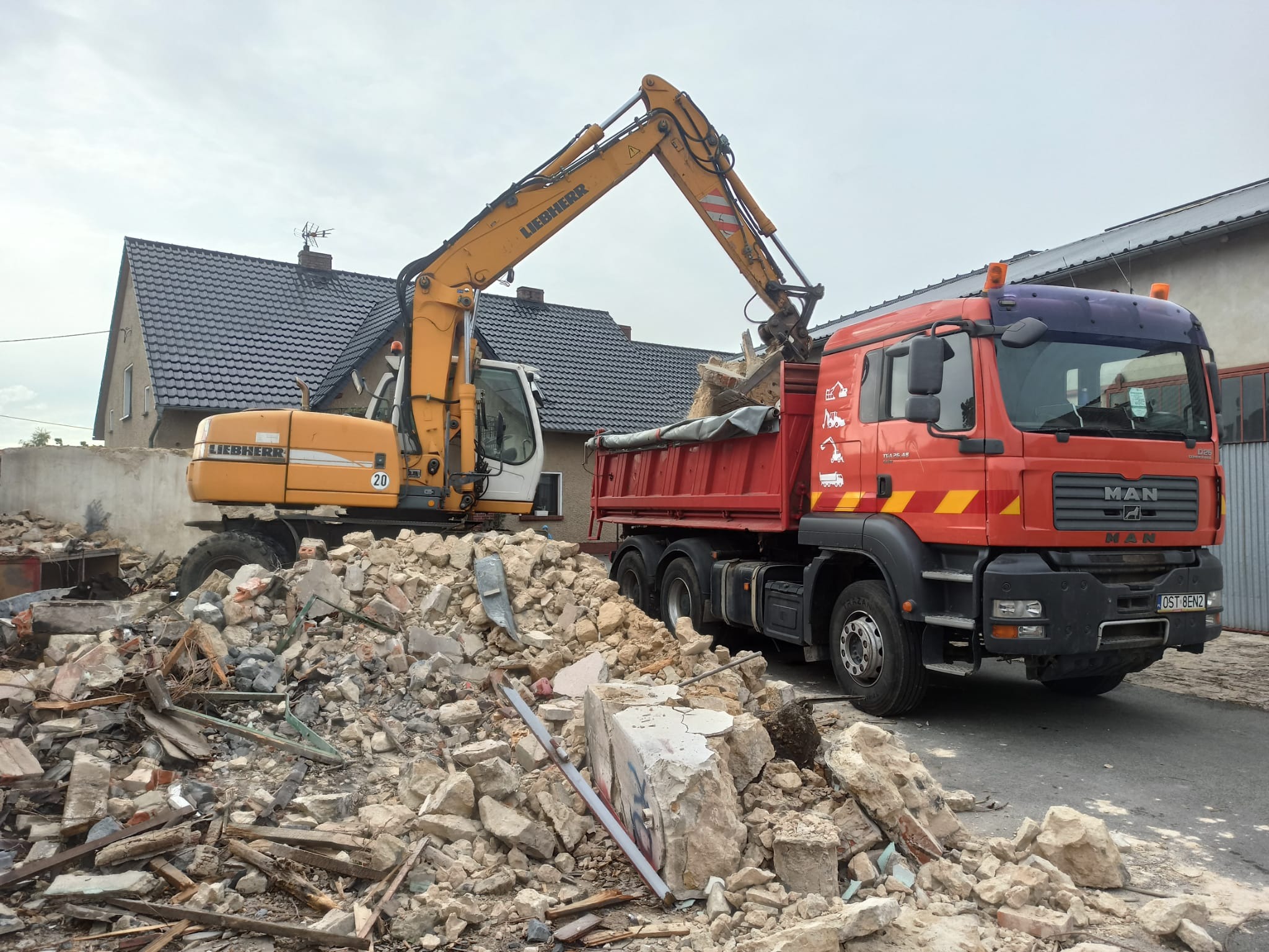 Żółta koparka Liebherr ładuje gruz do czerwonej ciężarówki MAN na tle rozbieranego budynku. Widoczny fragment dachu i sterta gruzu.